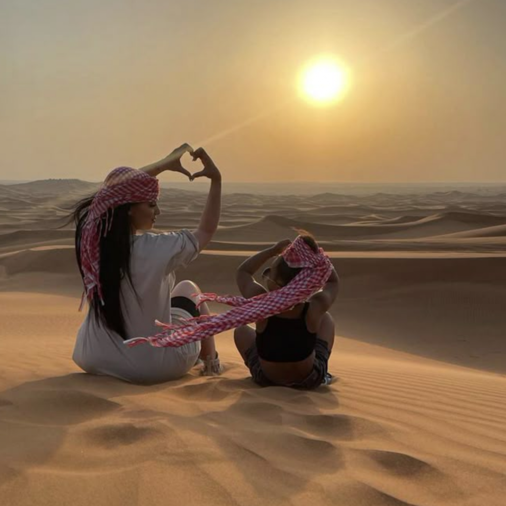 Mère et enfant assis dans les dunes de Chegaga au coucher du soleil, portant un keffieh rose, formant un cœur avec les mains.