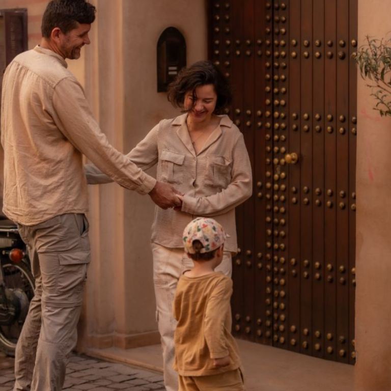 Famille heureuse devant la porte d’un riad traditionnel à Ouarzazate, en tenue claire, partageant un moment complice.