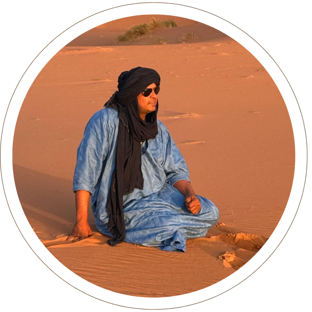 Mohamed Sbai assis dans le désert de M’Hamid, en tenue traditionnelle bleue et chèche noir