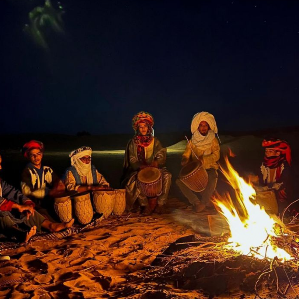 Musiciens nomades jouant du tambour autour d’un feu de camp dans le désert marocain, sous un ciel étoilé.